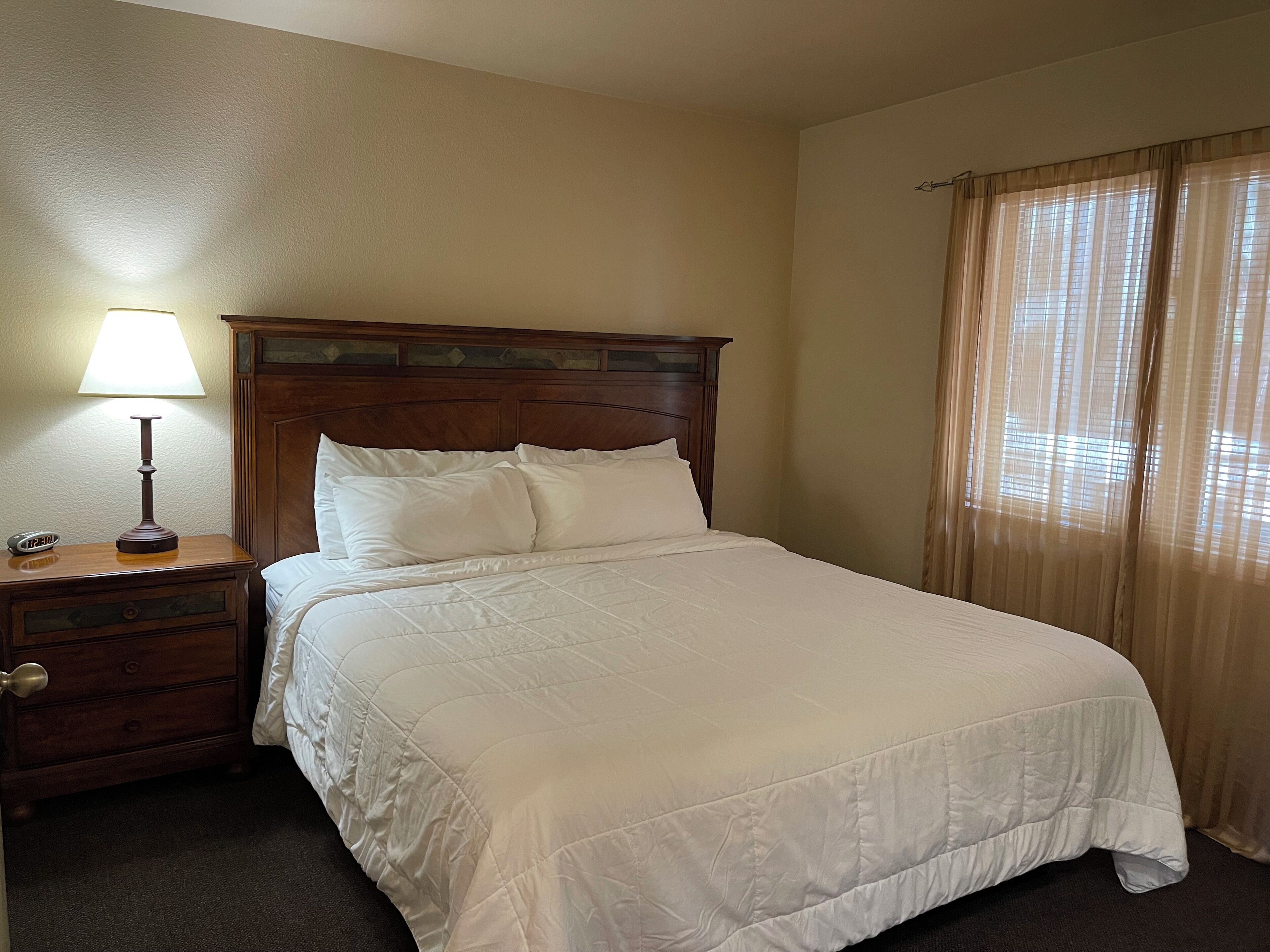 A neatly made bed with a wooden headboard, a bedside table with a lamp, and a window with sheer curtains.