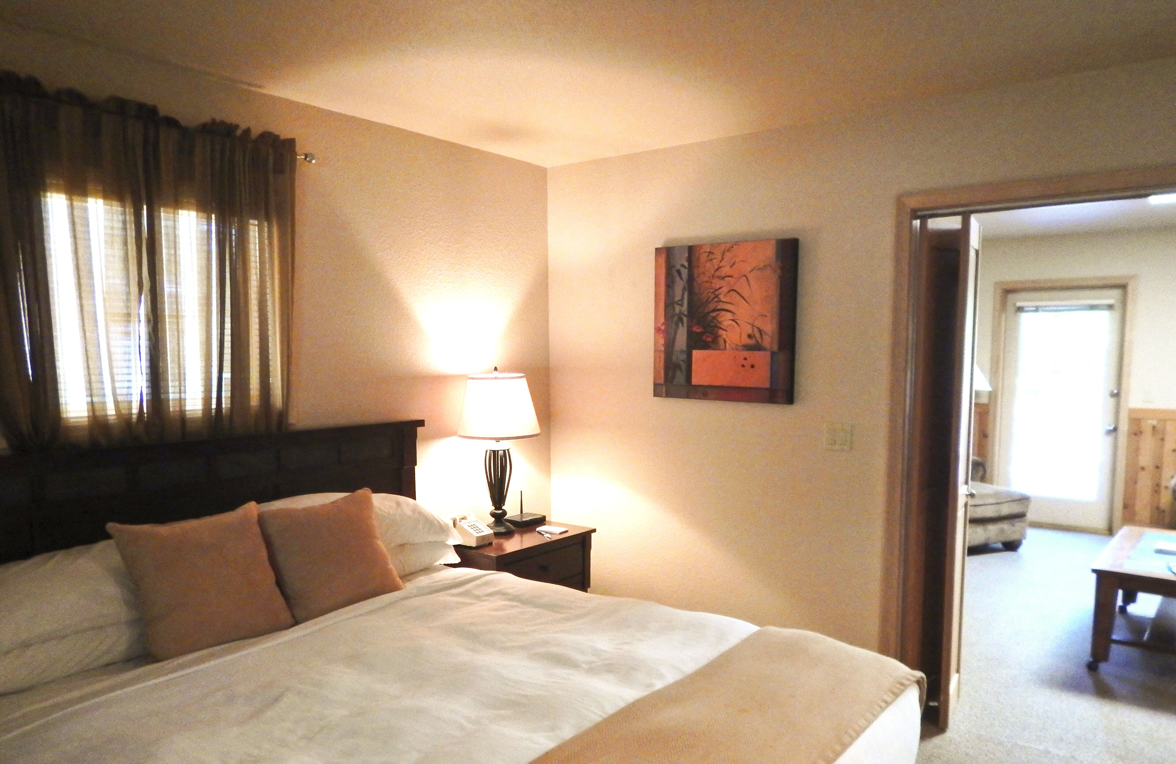 A hotel room with a bed, a nightstand with a lamp, a painting on the wall, and a door leading to another room.