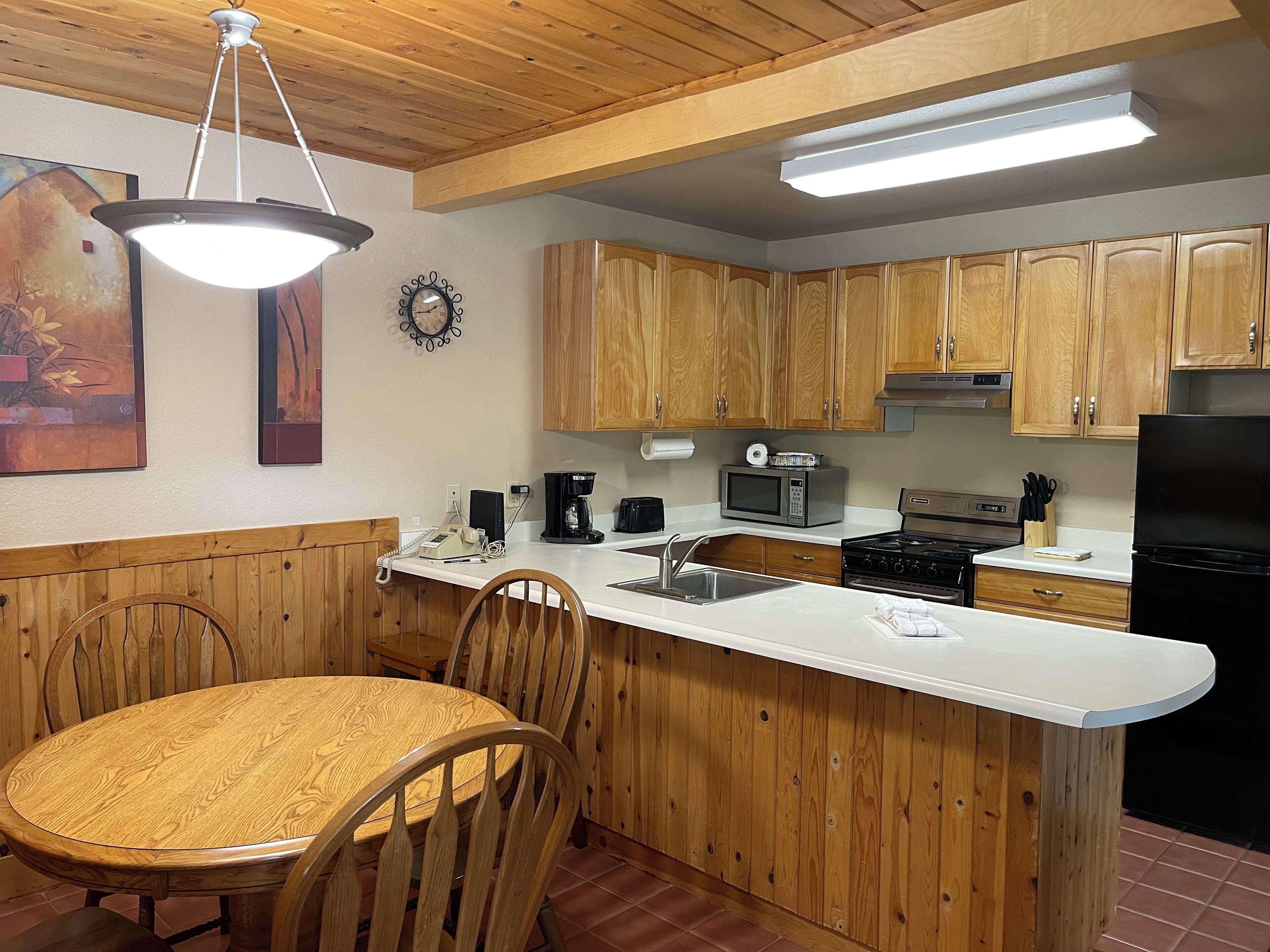 A kitchen with wooden cabinets, a white countertop, a dining table with chairs, a coffee maker, a microwave, a stove, and a refrigerator.