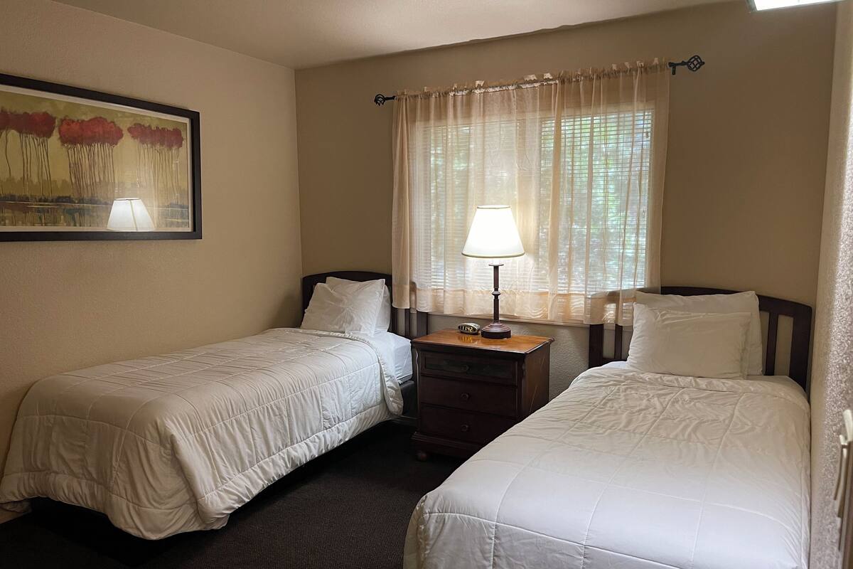 A hotel room with two single beds, a nightstand with a lamp, a painting on the wall, and a window with sheer curtains.
