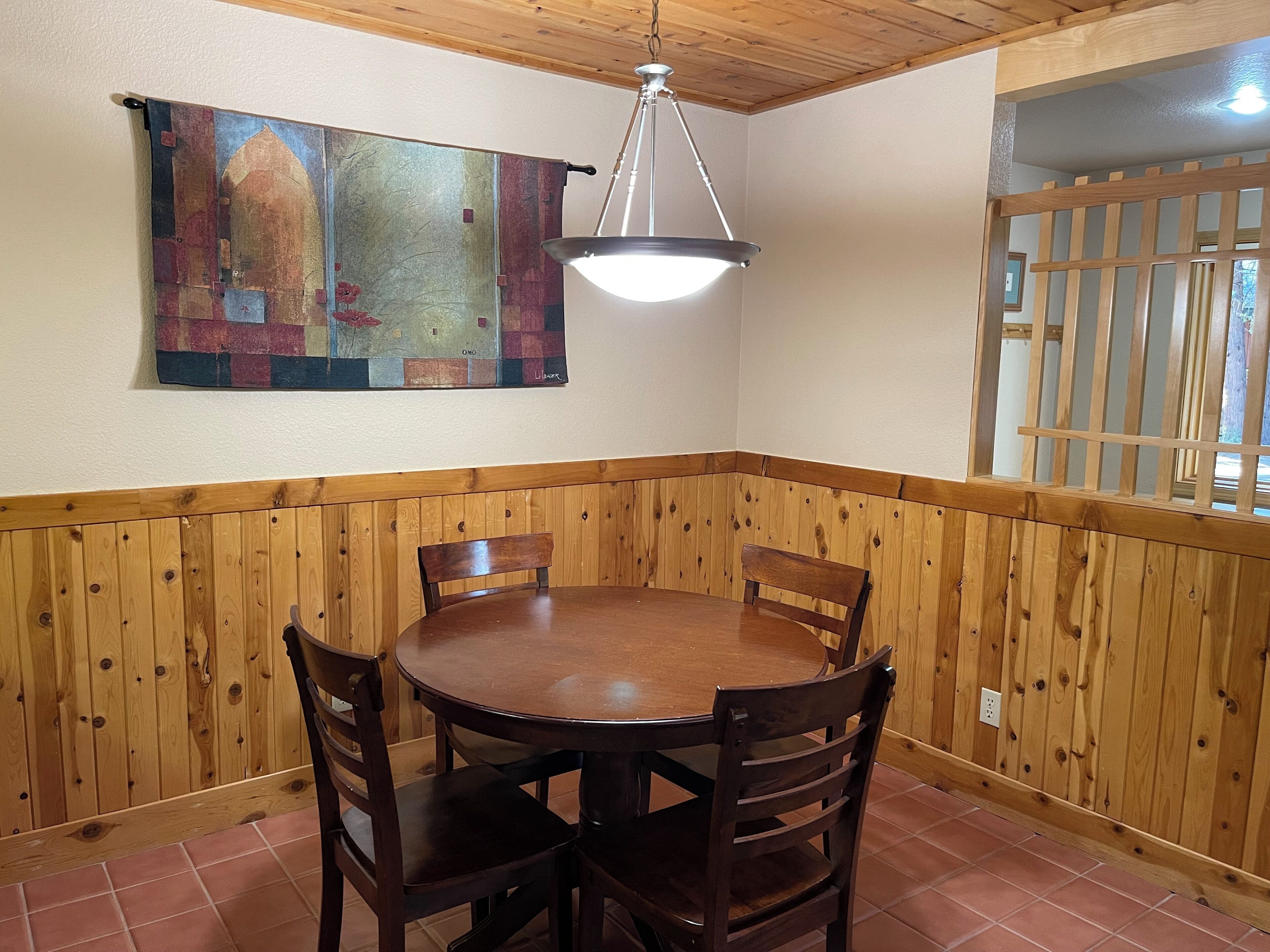 Un coin repas avec une table et des chaises en bois, un luminaire suspendu et une œuvre d’art colorée accrochée au mur.