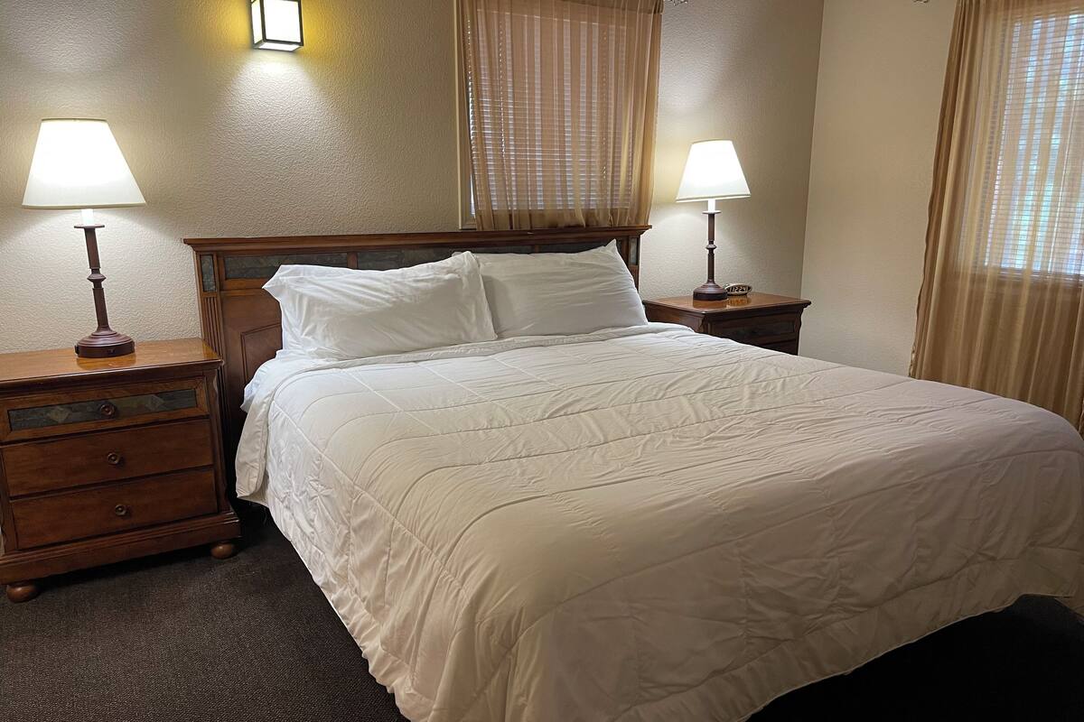 A neatly made bed with white bedding in a hotel room. There are two bedside tables with lamps, a wooden headboard, and a window with blinds.
