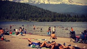 Una playa con gente tomando el sol y nadando, una montaña al fondo y una zona boscosa.