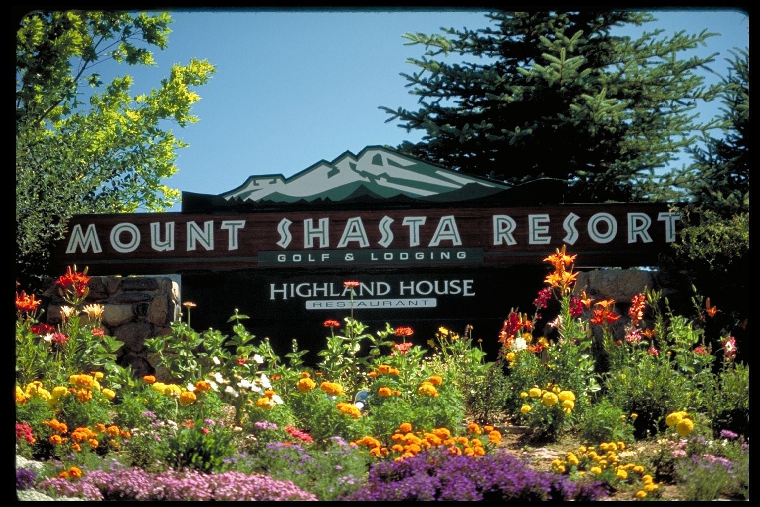 Schild für das Mount Shasta Resort, mit Bergpanorama und einem Garten voller bunter Blumen im Vordergrund.
