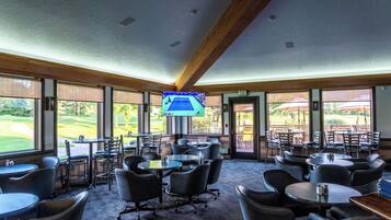 A spacious indoor area with multiple tables and chairs, large windows offering views of a golf course, and a television mounted on the ceiling.