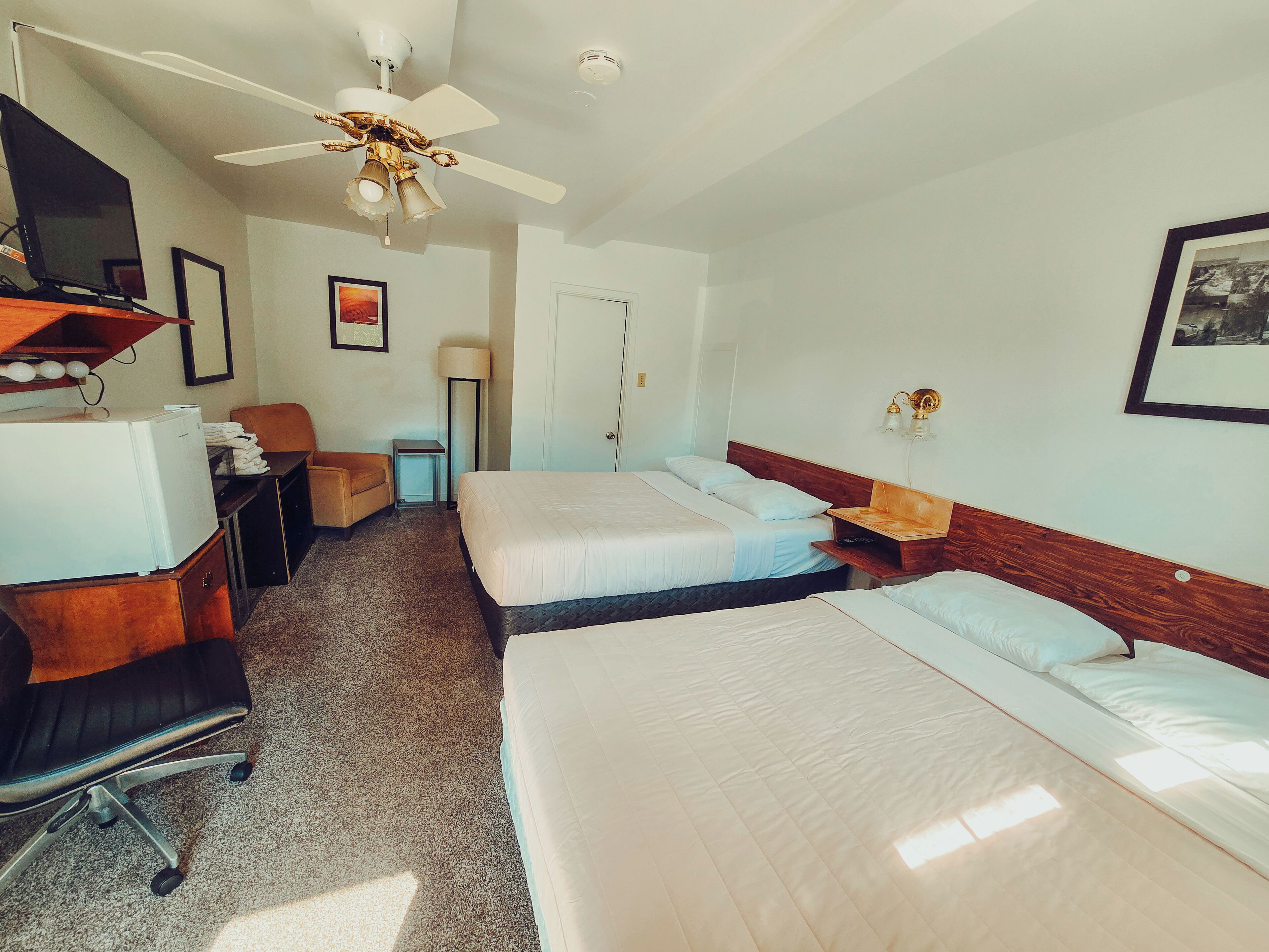 Ein Hotelzimmer mit zwei Betten, einem Schreibtisch mit Computer, einem Stuhl, einem Deckenventilator und einer Lampe.