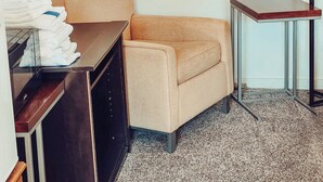 Une chambre d’hôtel avec un fauteuil beige, une petite table avec une lampe, un tableau encadré au mur et une pile de serviettes pliées sur une commode.