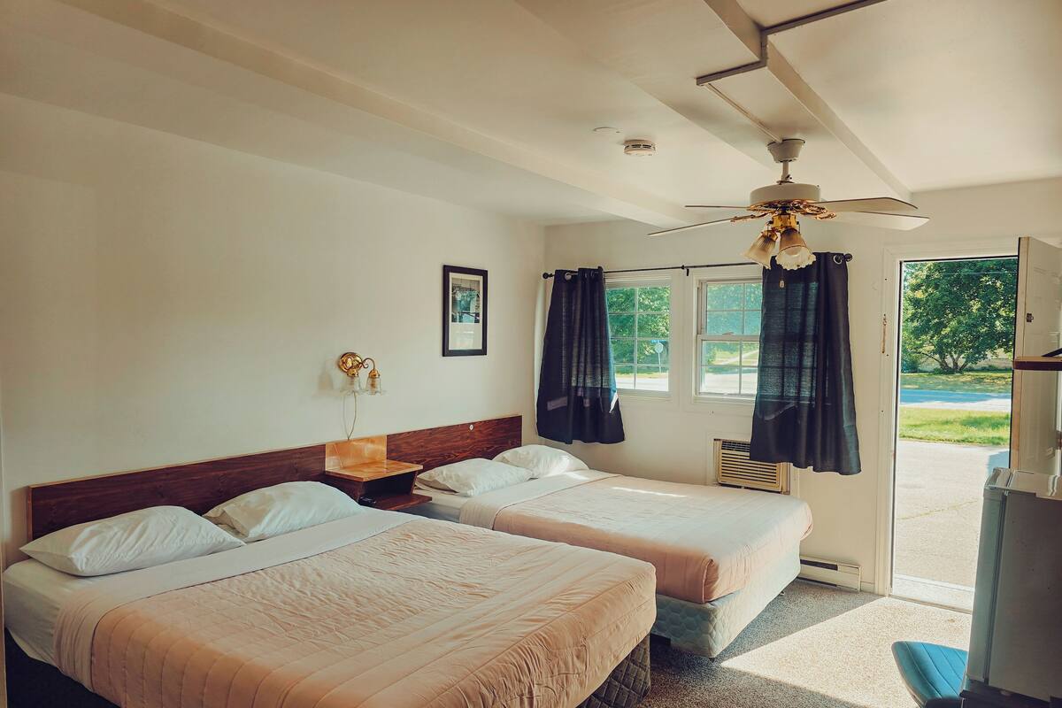 Habitación de hotel con dos camas, ventilador de techo, ventana con cortinas, un cuadro en la pared y vistas al exterior.