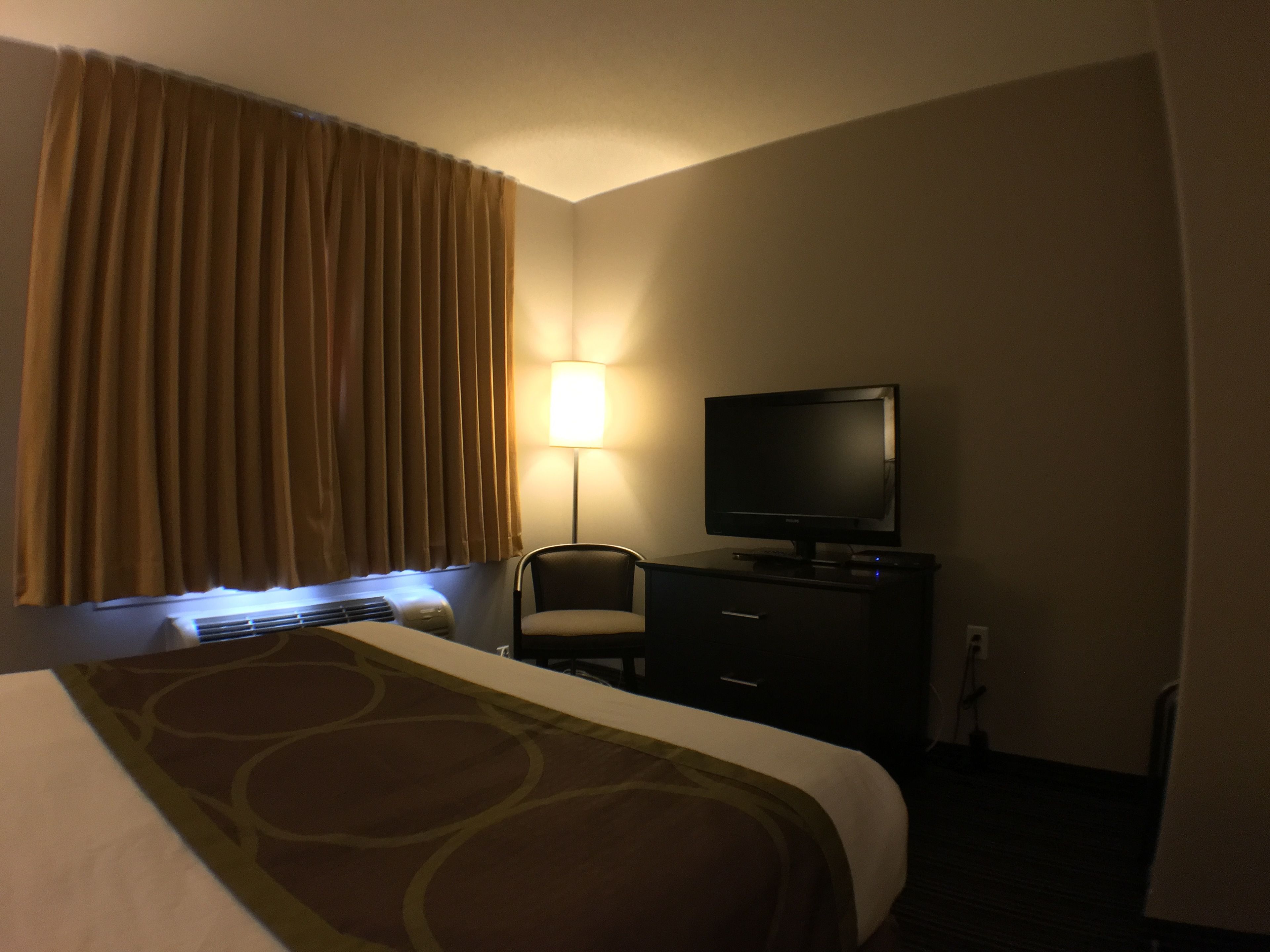 A hotel room with a bed, a television on a dresser, a lamp, and a window with curtains.
