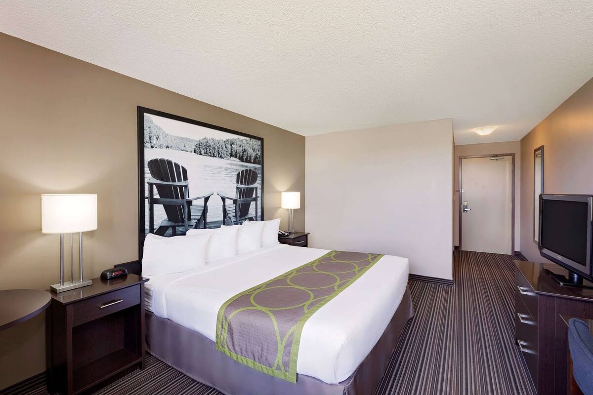 Quarto de hotel com uma cama grande, duas mesas de cabeceira com luminárias, uma TV de tela plana e um quadro grande na parede.