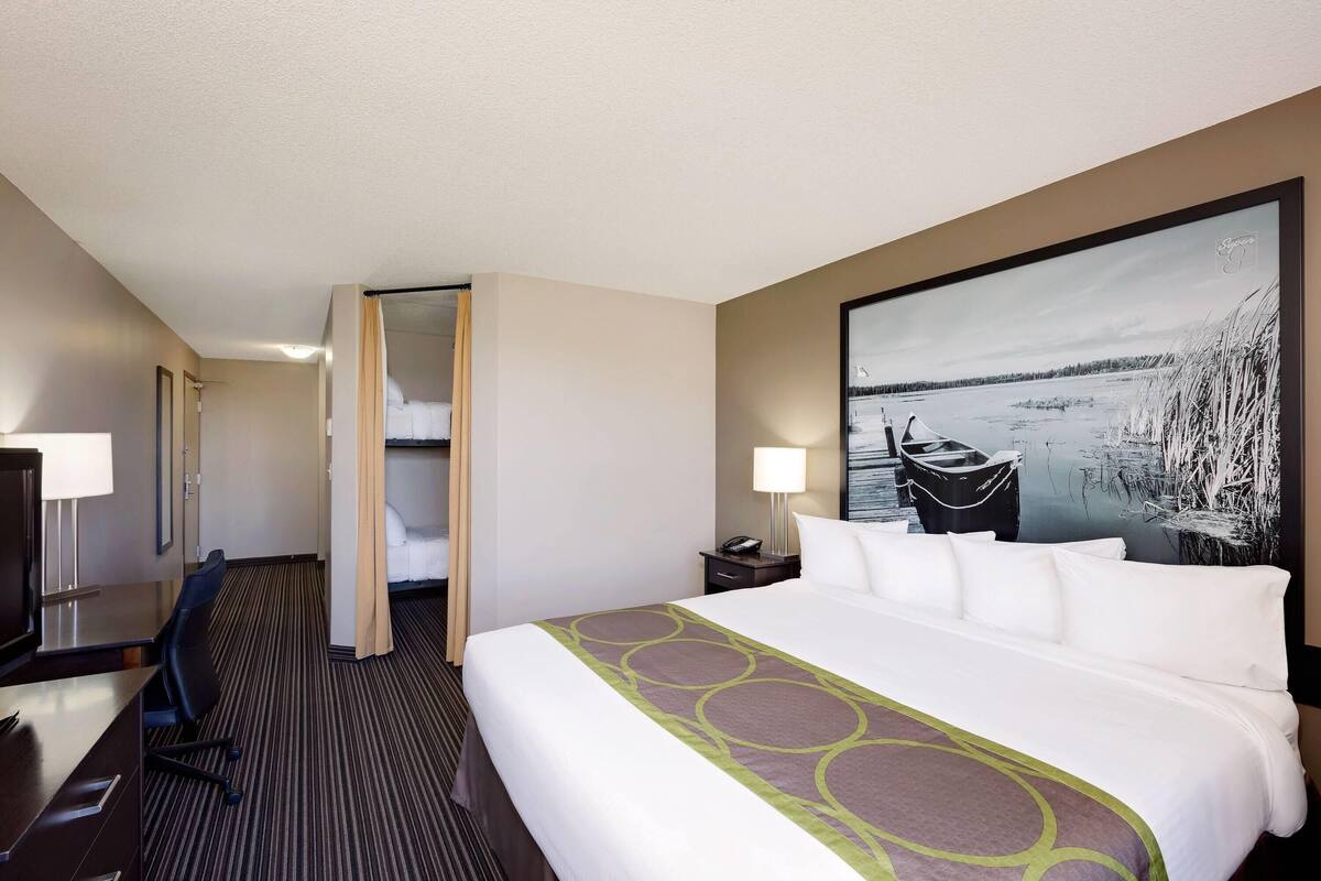 Quarto de hotel com cama, escrivaninha, cadeira, mesa de cabeceira, abajur e uma grande pintura de uma paisagem lacustre.