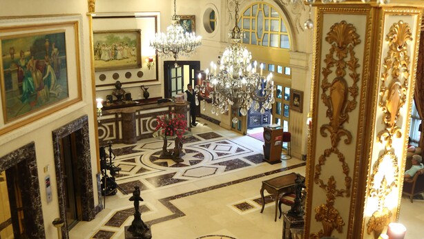 Un lobby espacioso con candelabros ornamentales, piso de mármol y una escalera que conduce a los pisos superiores.