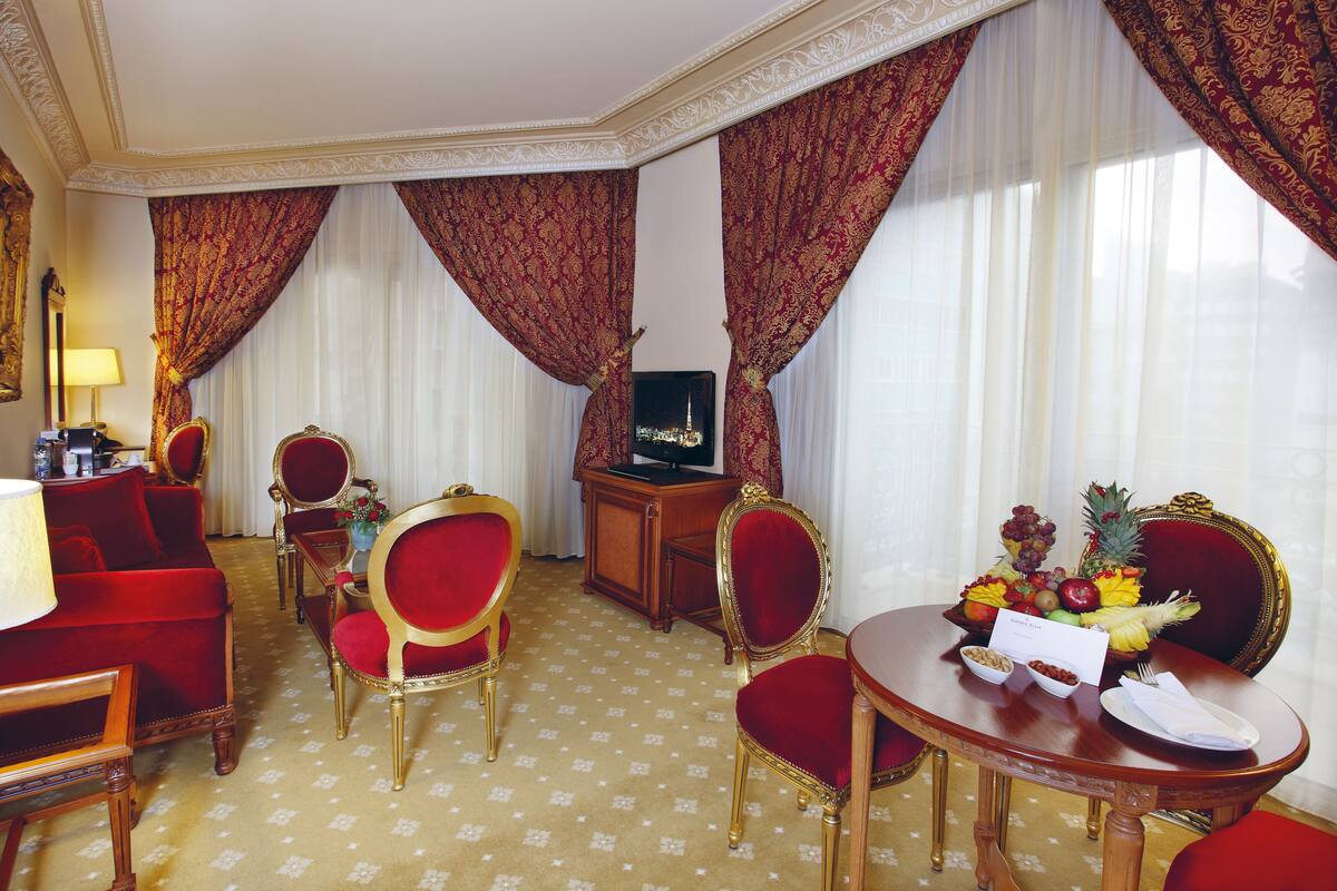 A hotel room with a red sofa, two red chairs, a wooden table with fruit and a menu, and a television on a wooden cabinet.