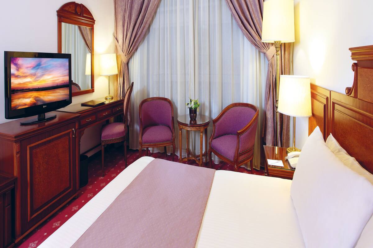 A hotel room with a bed, two armchairs, a desk with a TV, and a small table with a lamp.