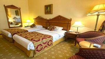 A hotel room with two beds, a sitting area with a chair, a bedside table with a lamp, and a large mirror on the wall.