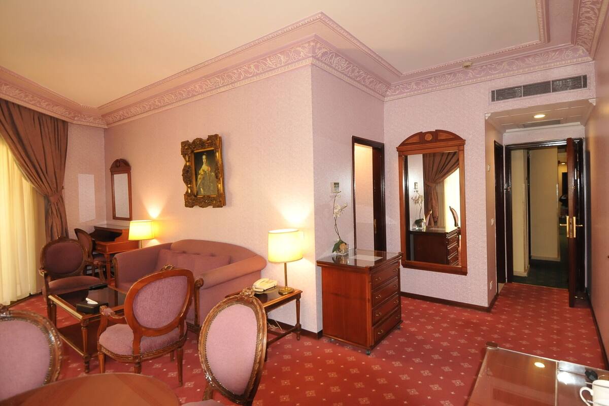 A hotel room with a red carpet, a sofa, a chair, a dresser, a mirror, and a door leading to another room.