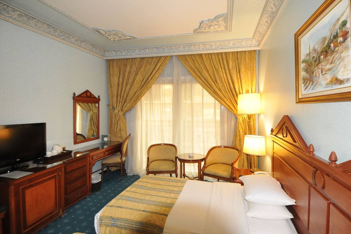 A hotel room with a bed, a desk with a television, two armchairs, a small table, and a painting on the wall.