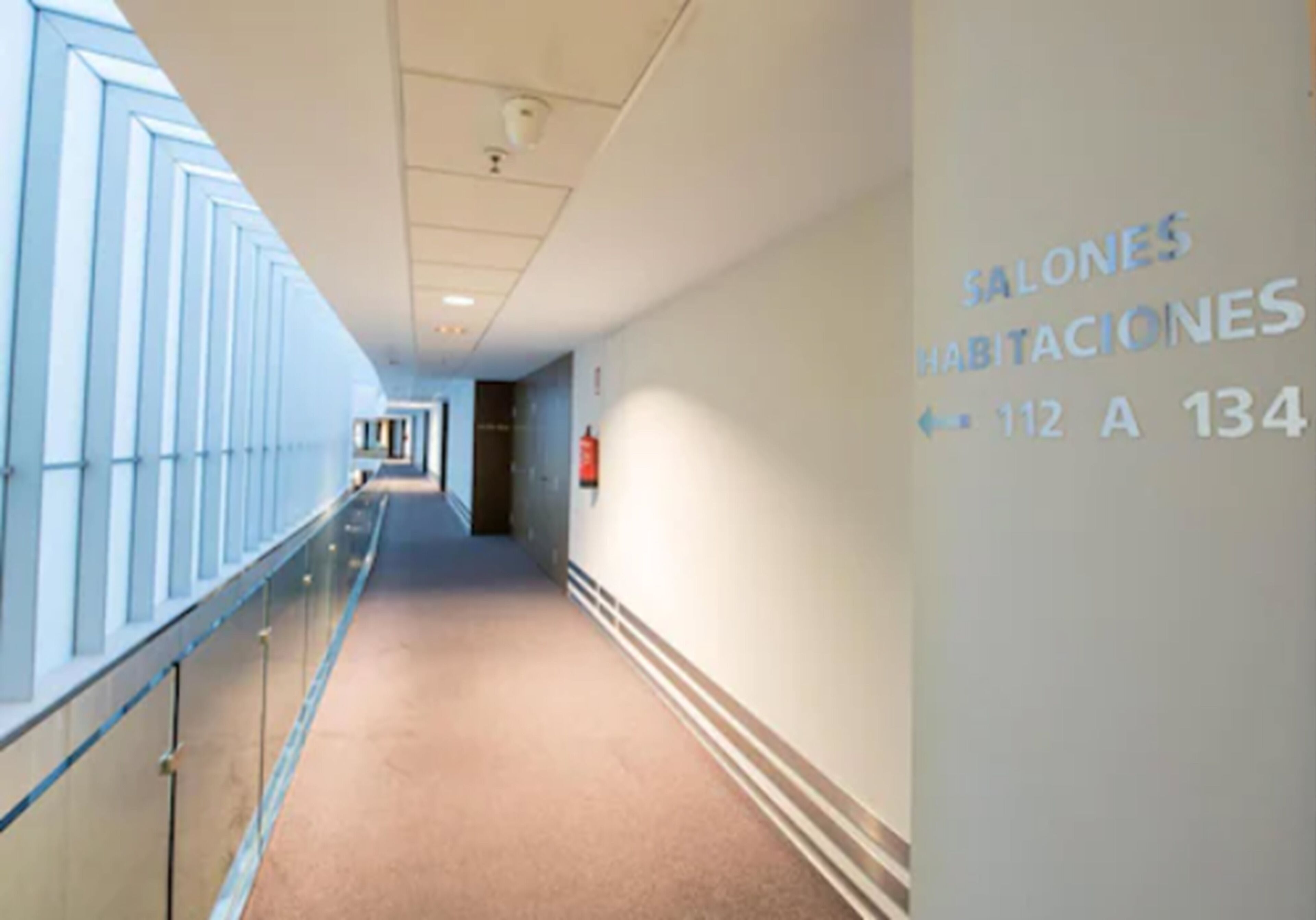 Un pasillo de hotel con piso alfombrado, barandales de vidrio y una pared con la inscripción 'SALONES HABITACIONES' y los números de habitación 112 a 134.