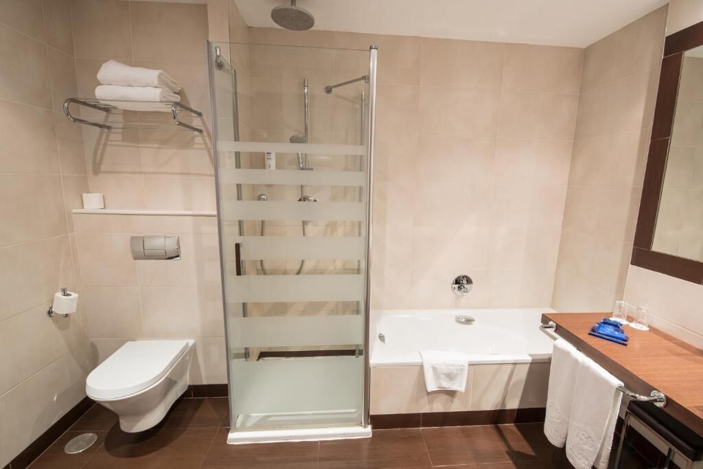 A modern bathroom with a glass-enclosed shower, a white toilet, a bathtub, and a wooden vanity with a towel rack.