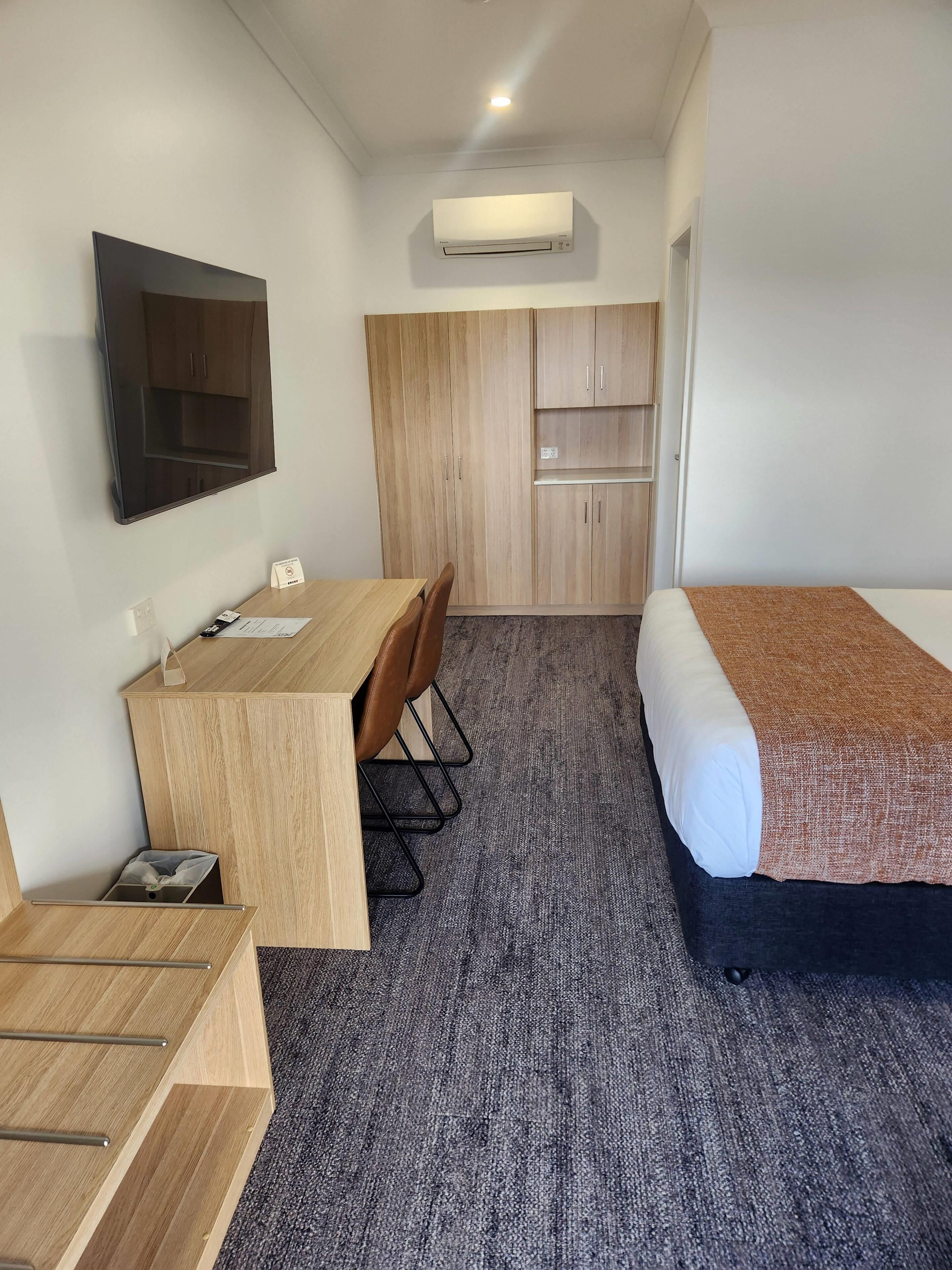 A hotel room with a bed, a desk with a chair, a TV mounted on the wall, and a wardrobe.