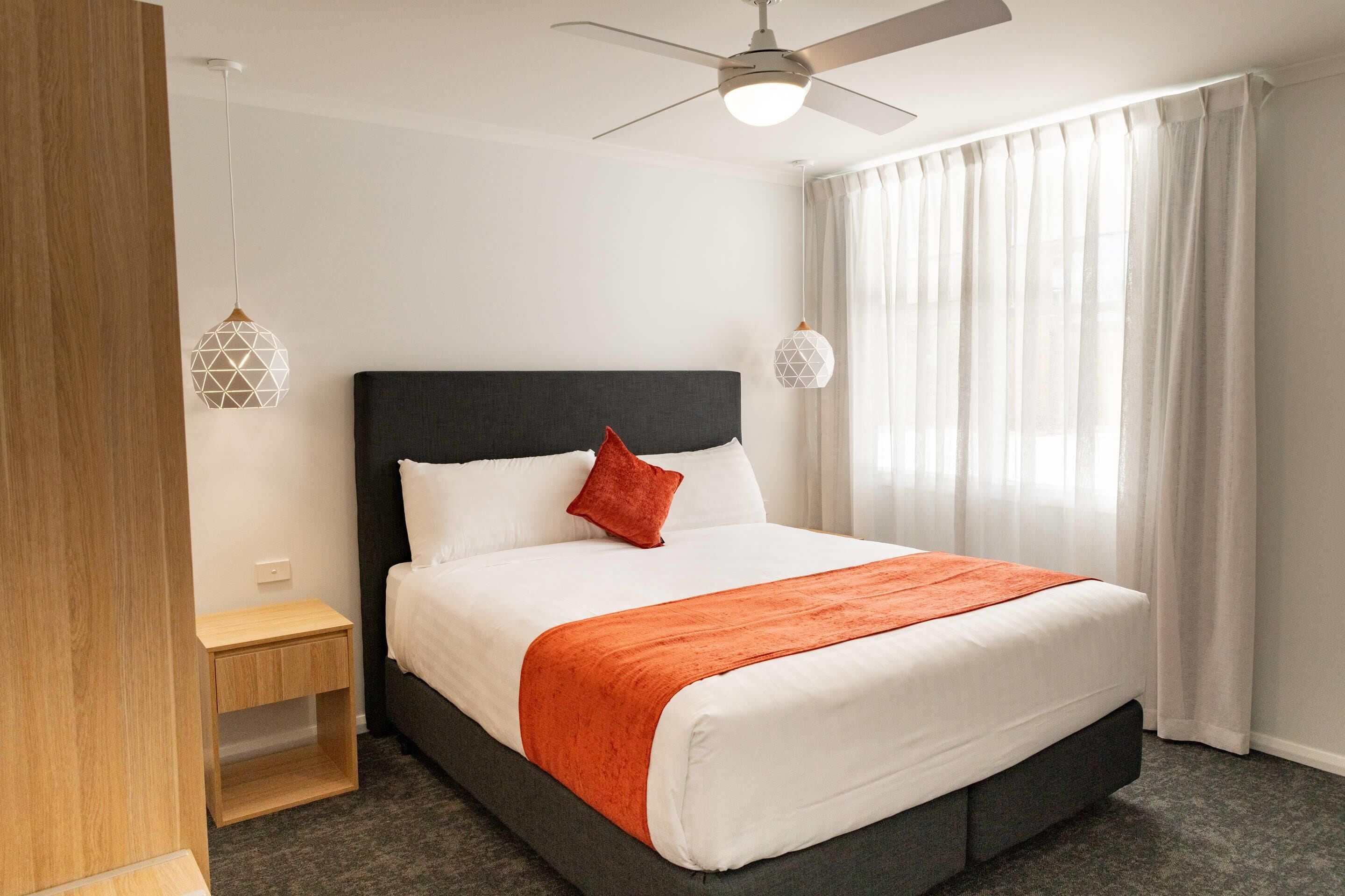 Quarto de hotel com cama, mesa de cabeceira, ventoinha no teto e janela com cortinas.