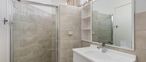 A bathroom with a glass shower enclosure, a white sink, and a mirror with a shelf above it.