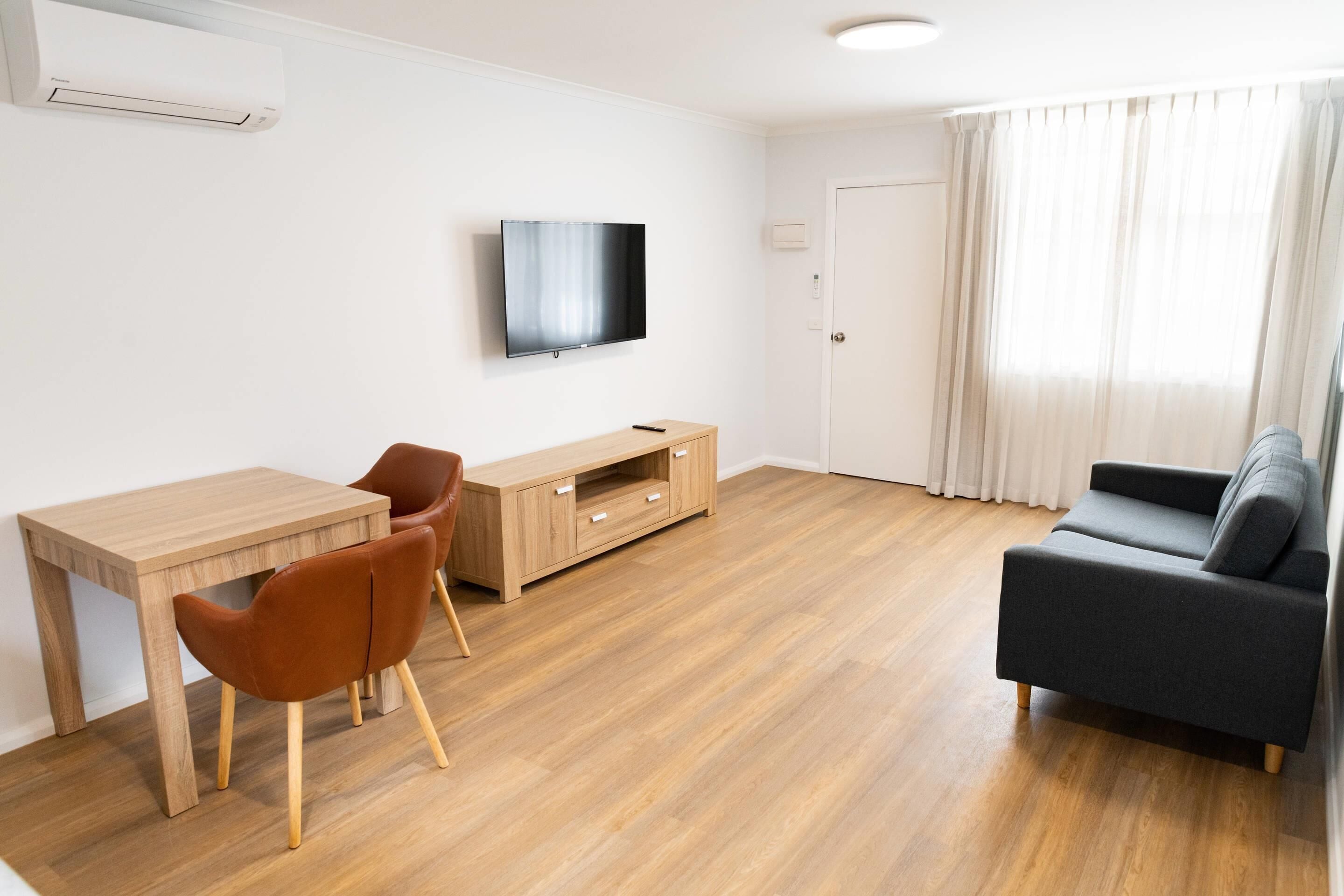 A modern living room with a wooden floor, a grey sofa, a wooden coffee table, and a TV mounted on the wall.