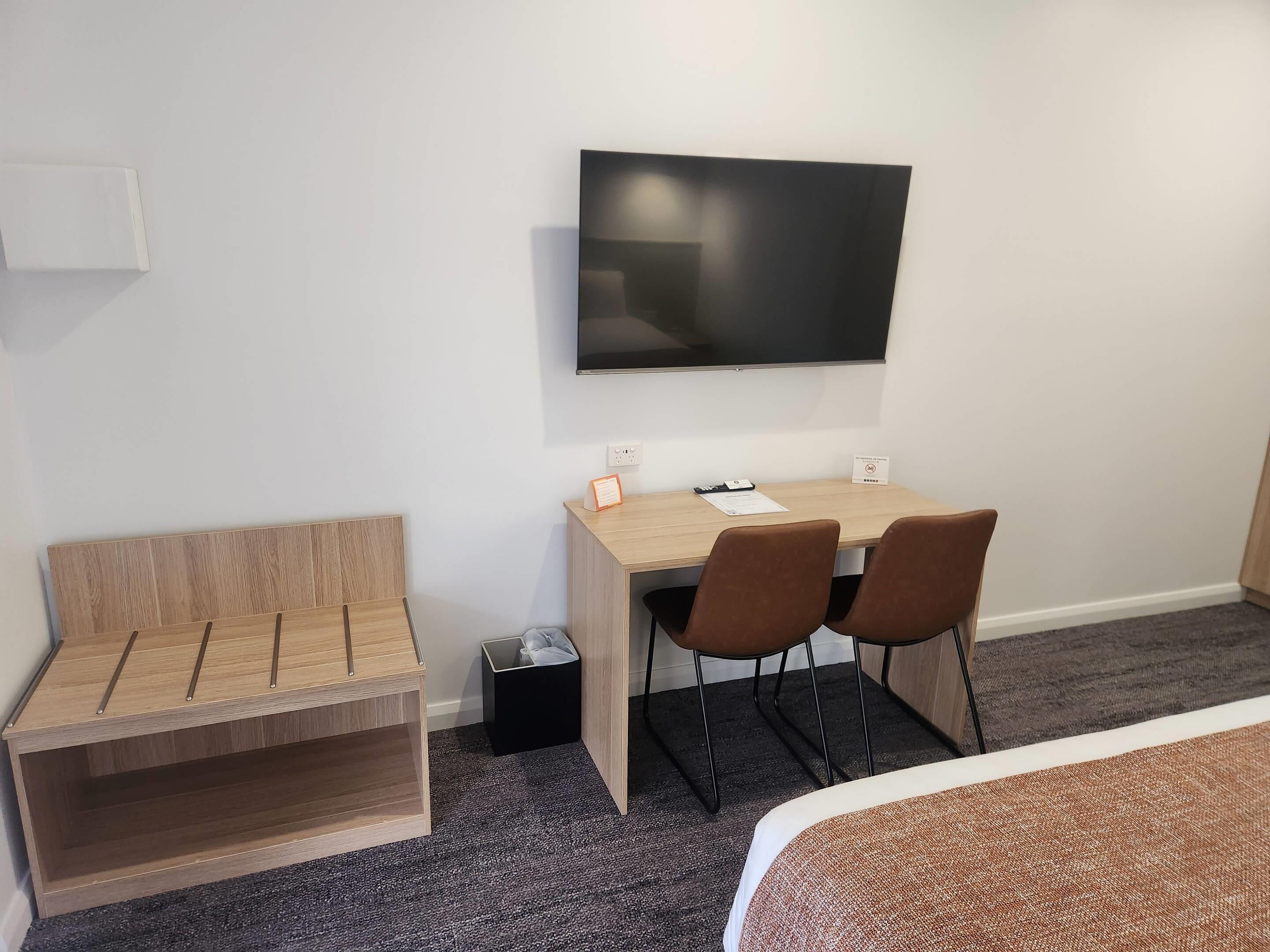 Habitación de hotel con escritorio de madera, dos sillas, un televisor montado en la pared, un cubo de basura pequeño y una cama con cubre color marrón.