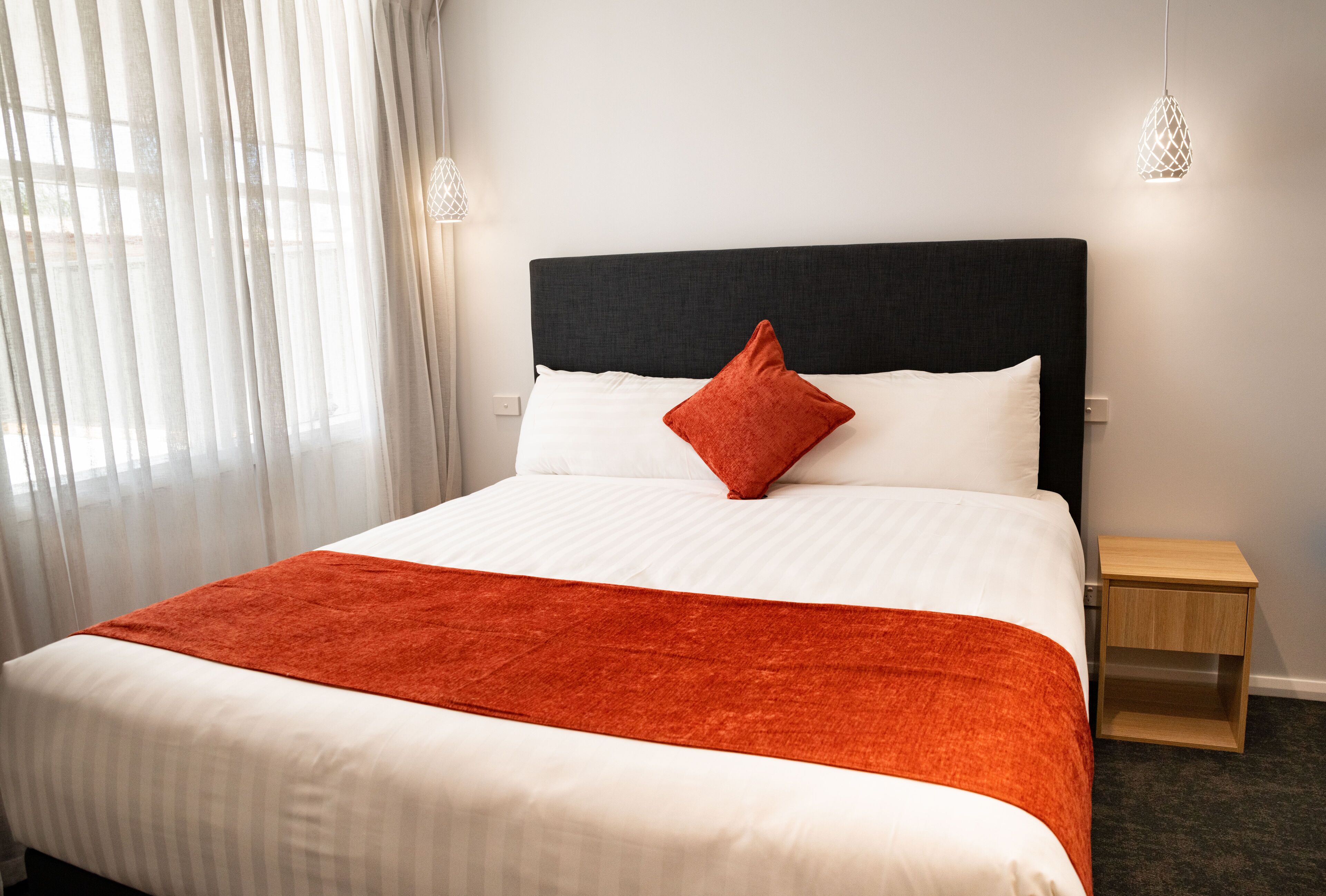 A neatly made bed with a white and orange bedspread, a black headboard, and a single red pillow. There is a small wooden nightstand beside the bed.