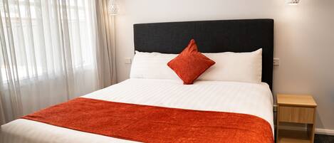 A neatly made bed with a white and orange bedspread, a black headboard, and a single red pillow. There is a small wooden nightstand beside the bed.