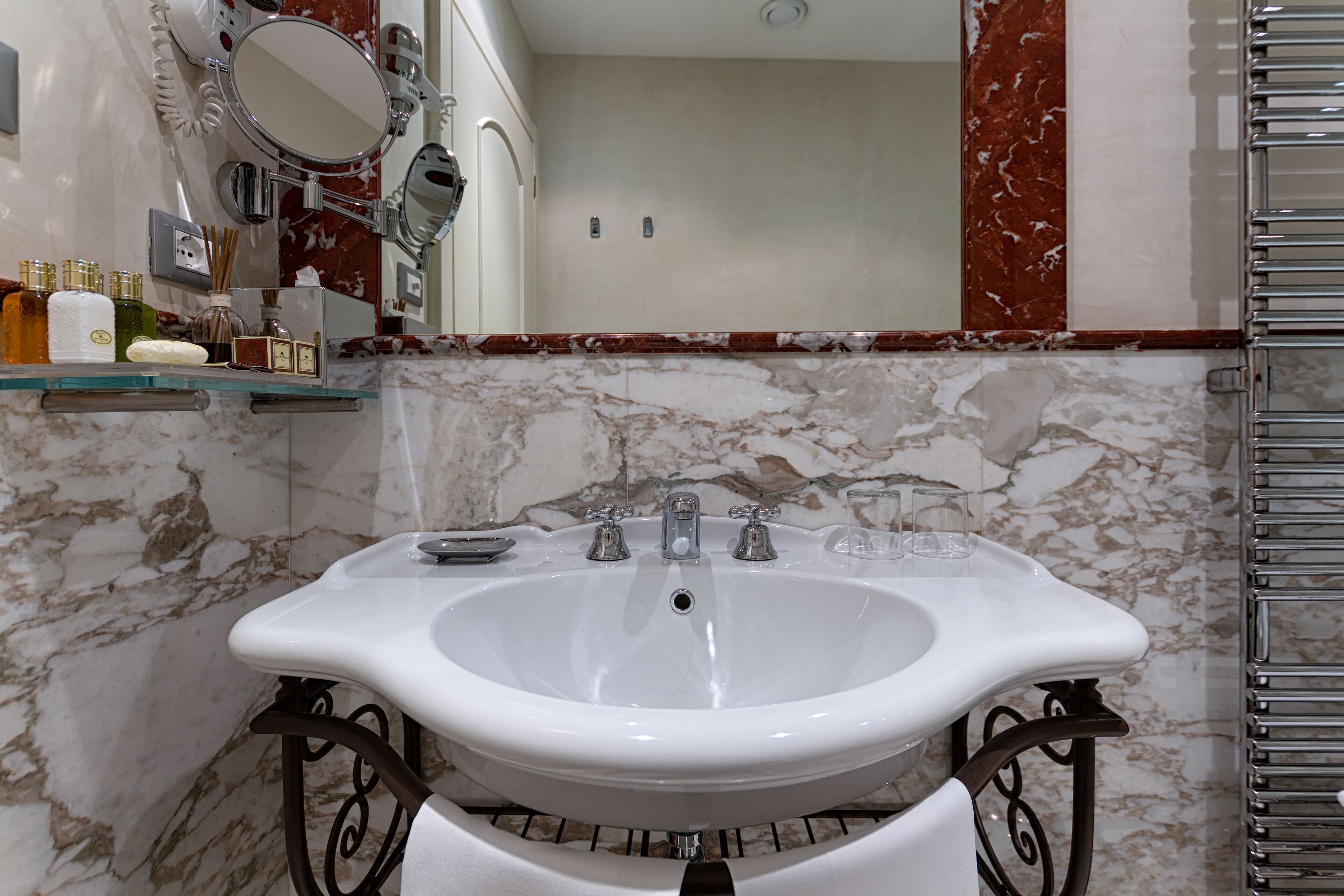 Une salle de bain avec un lavabo blanc, des murs en marbre et une étagère en verre où sont rangés les articles de toilette.