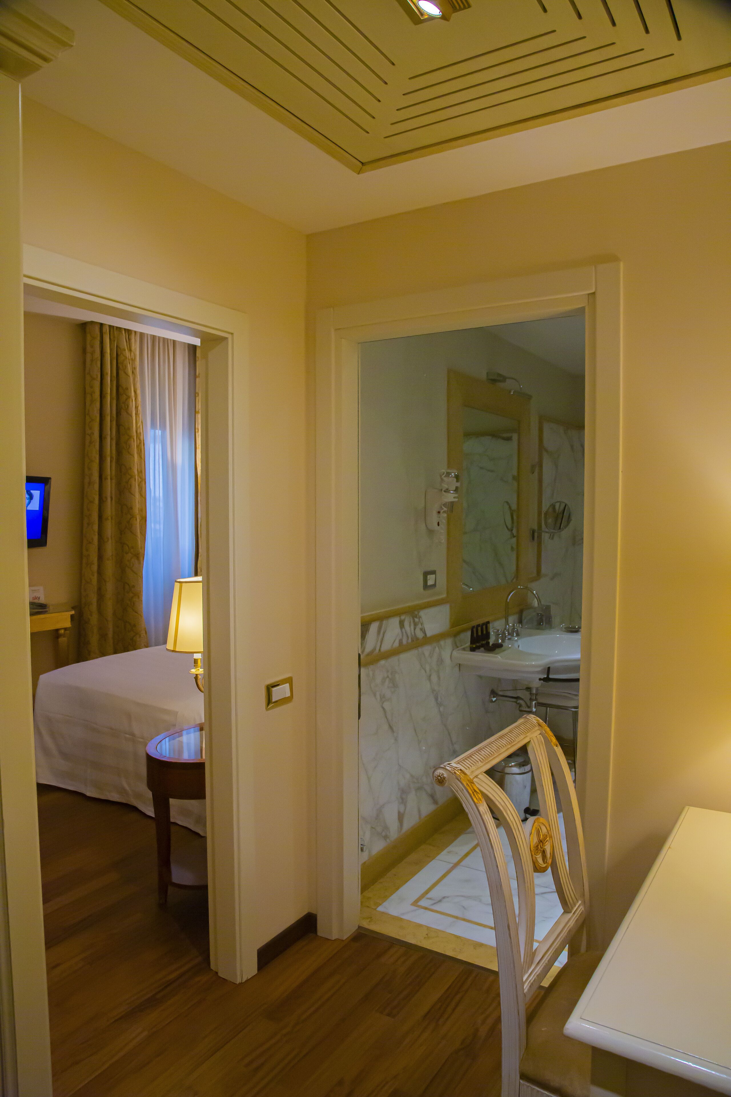 Une chambre d’hôtel comprenant un lit, une table de chevet avec une lampe, une salle de bain avec un miroir, un lavabo et une chaise.