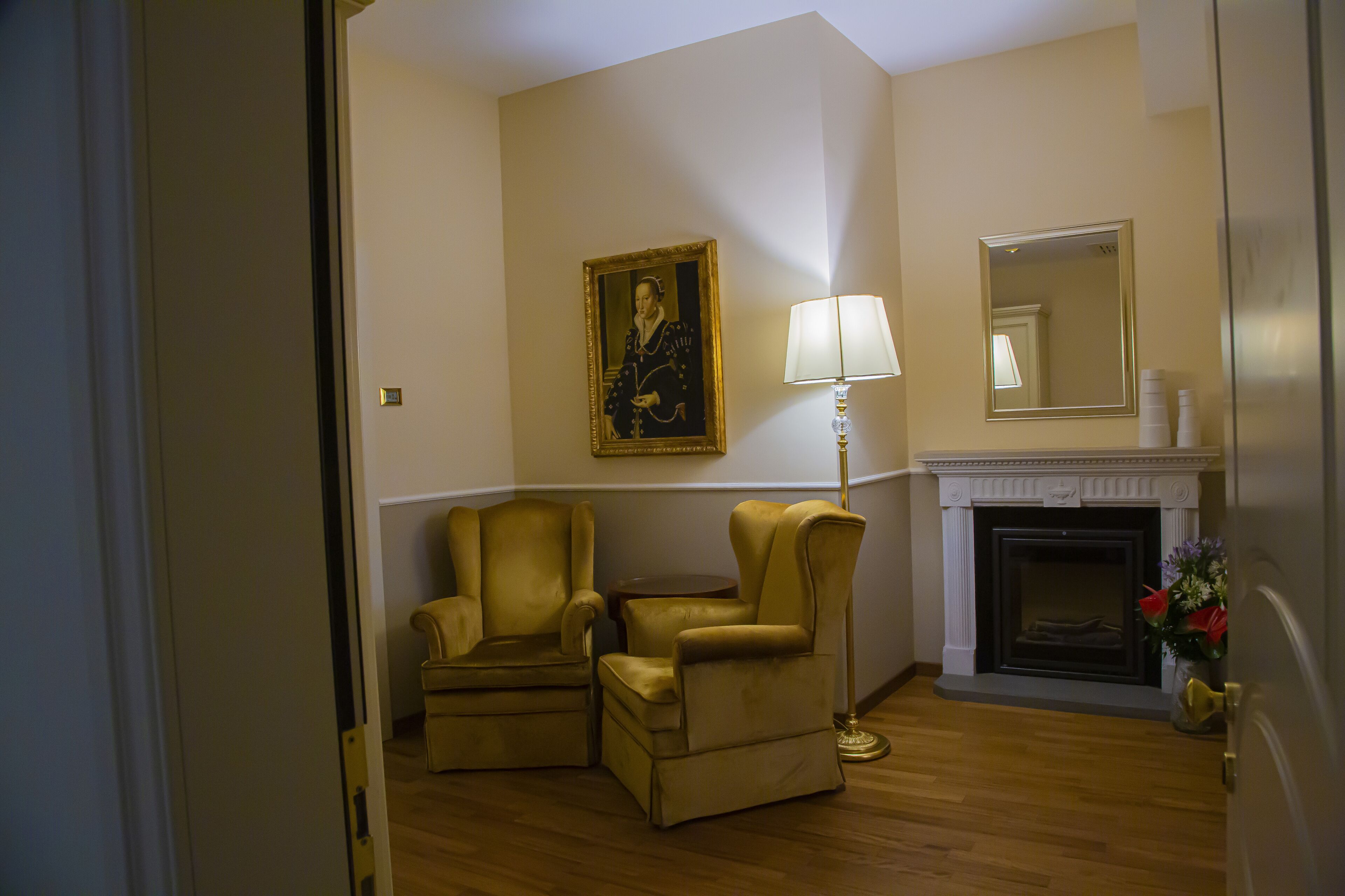 A room with two gold armchairs, a standing lamp, a fireplace, and a framed portrait on the wall.
