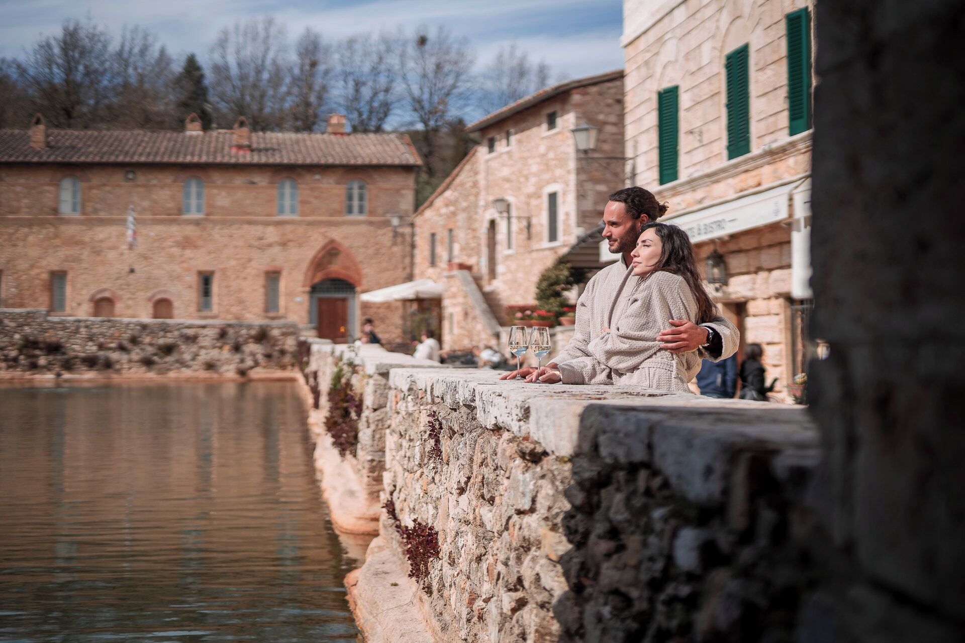 A couple standing by a stone wall overlooking a body of water, with historic stone buildings in the background.