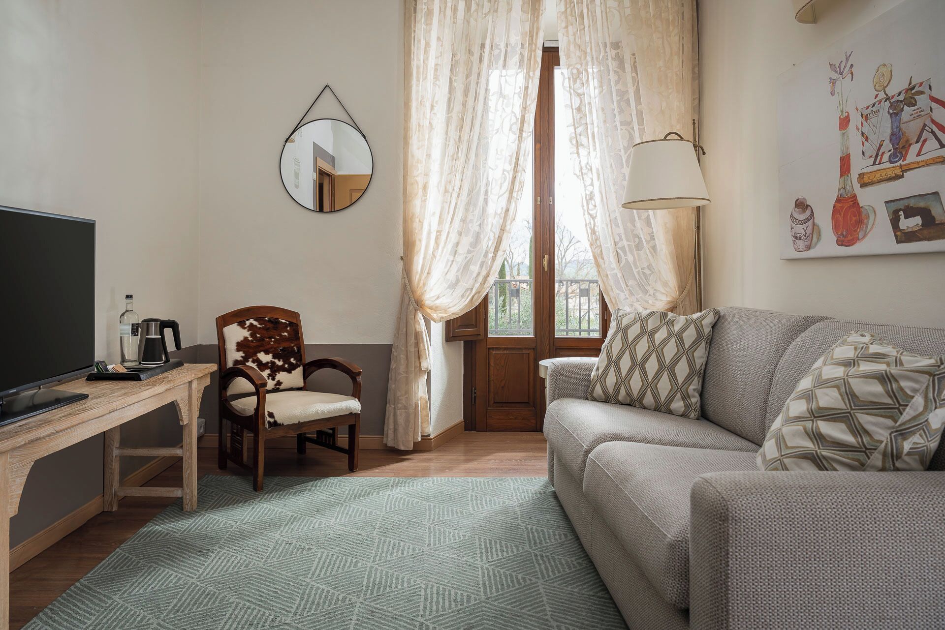 Un salon comprenant un bureau en bois, un fauteuil en cuir, un canapé, un miroir et une fenêtre ornée de rideaux légers.