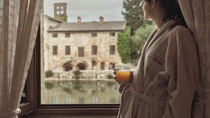 Una persona con bata sostiene un vaso de zumo de naranja, mirando por la ventana un edificio de piedra con un estanque delante.