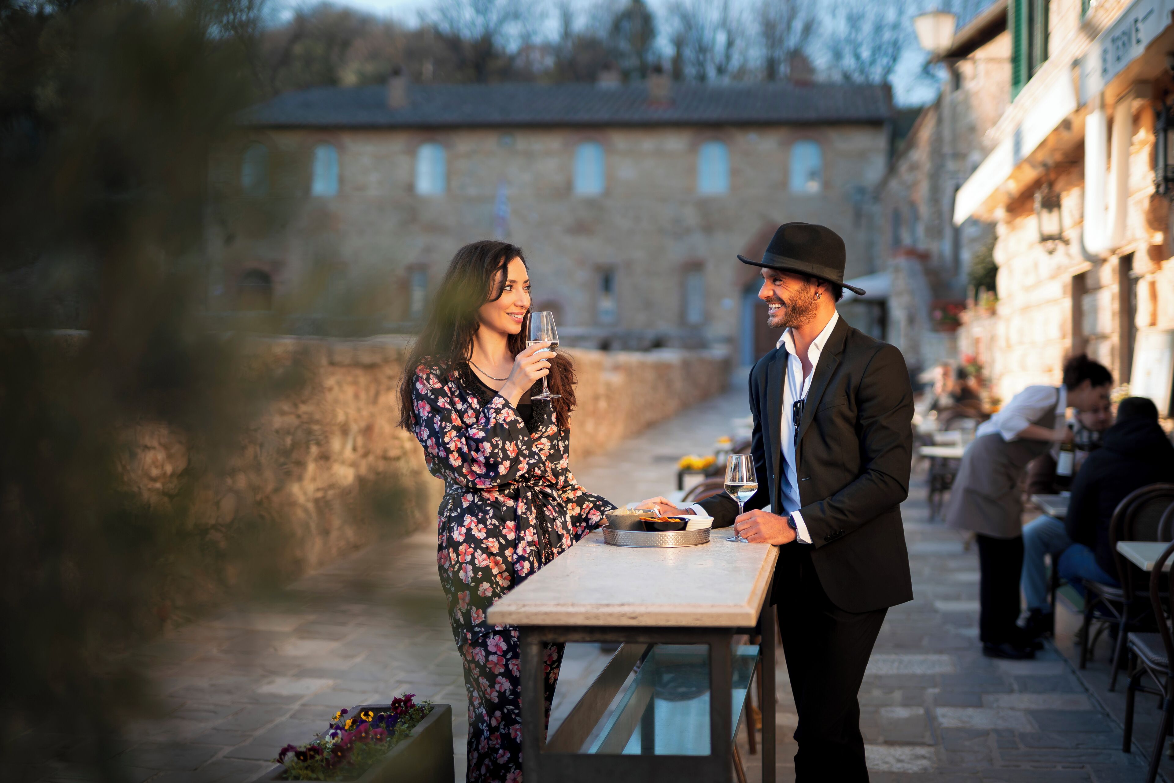 A couple dining at an outdoor table with a stone wall and buildings in the background.