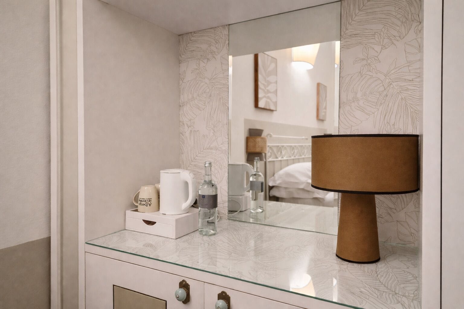 A modern bathroom with a glass countertop, a large mirror, a wooden lamp, and a bed visible in the reflection.