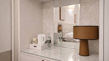 A modern bathroom with a glass countertop, a large mirror, a wooden lamp, and a bed visible in the reflection.