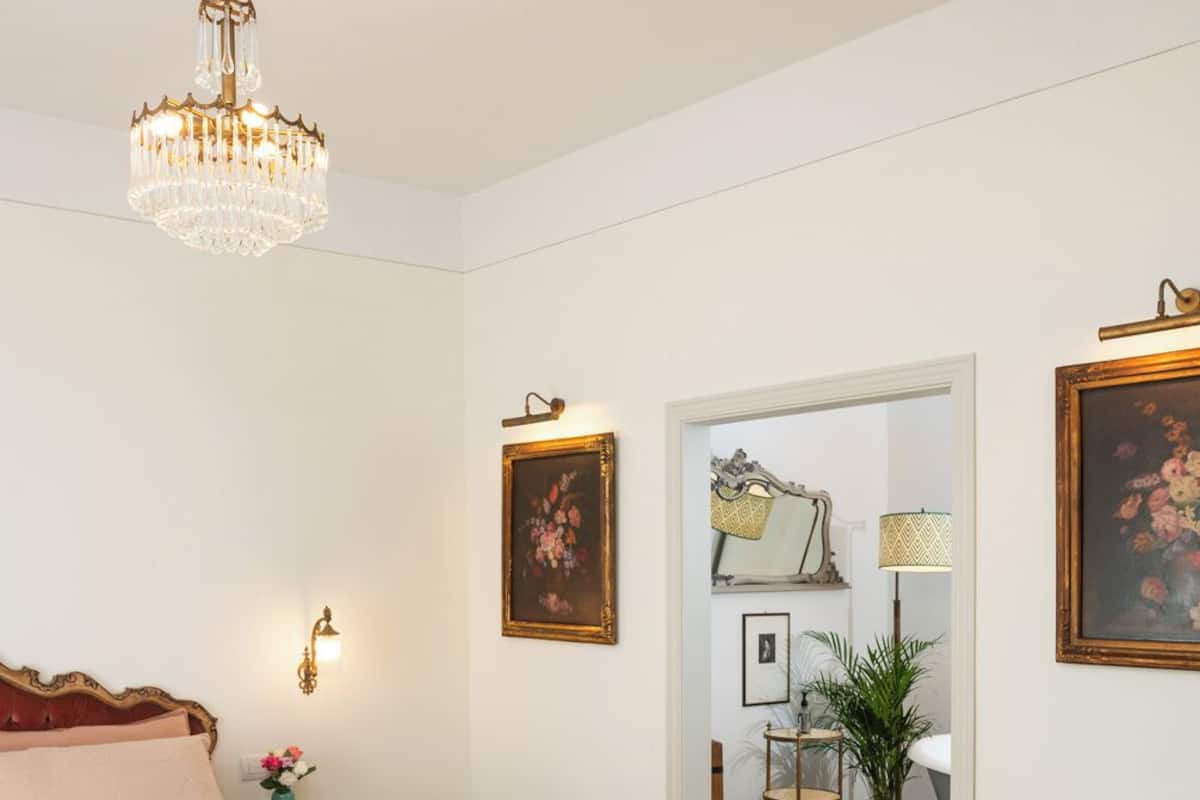Une chambre à coucher avec un lit, un plaid vert, un plancher en bois, un lustre, un bureau, une chaise et une porte donnant sur une autre pièce.