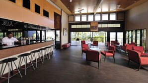 A bar area with stools, a counter, and a bartender. There are red chairs and tables outside with a view of greenery.