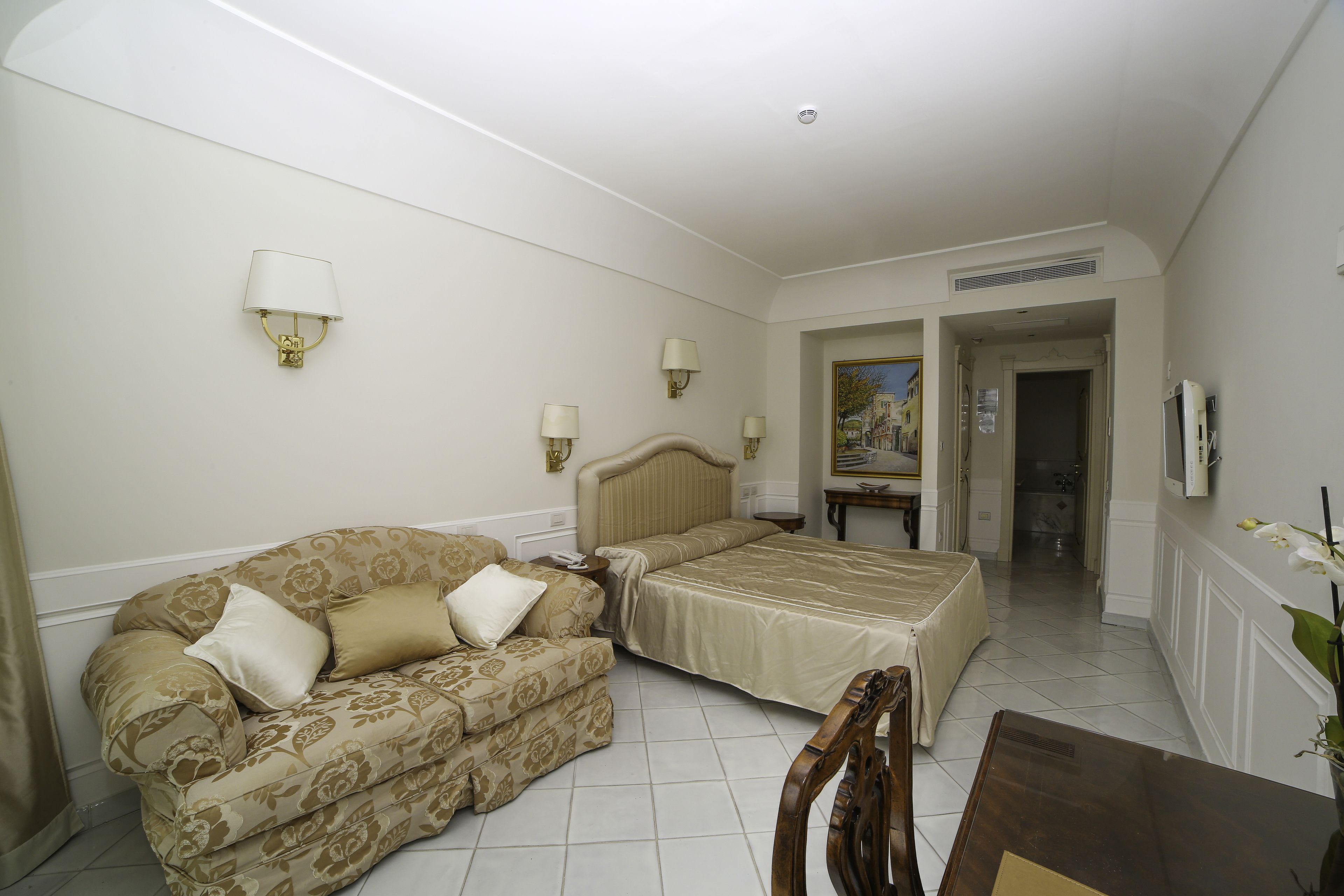 A hotel room with a bed, a sofa, a dining table, and two wall-mounted lamps.