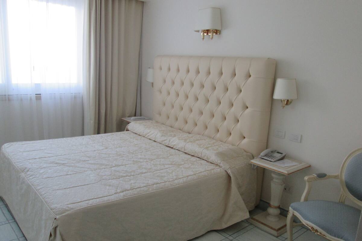 A hotel room with a large bed, a bedside table with a lamp, a chair, and a wall-mounted telephone.