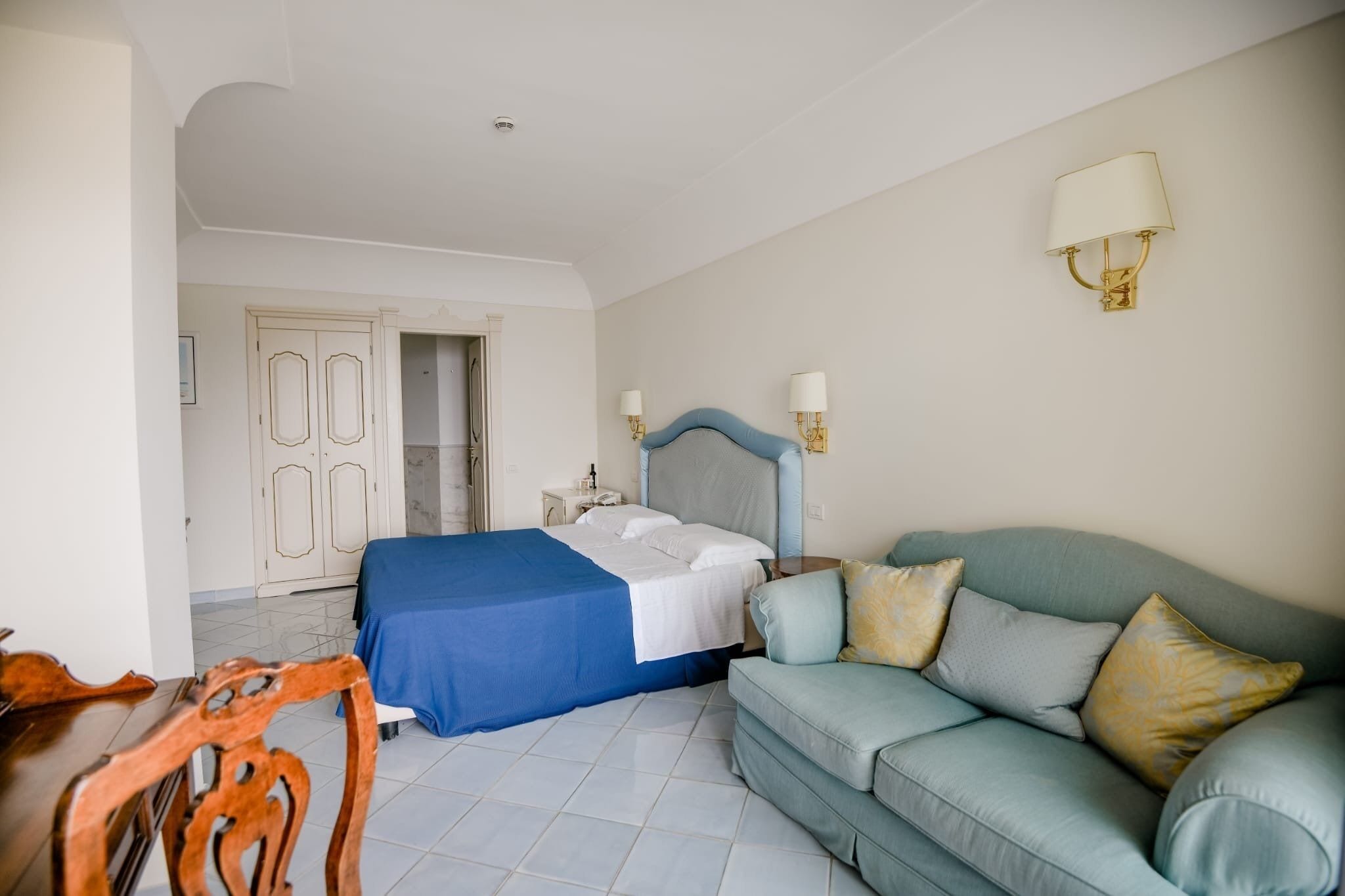 A hotel room with a bed, a sofa, a wooden chair, a desk, and a wardrobe.