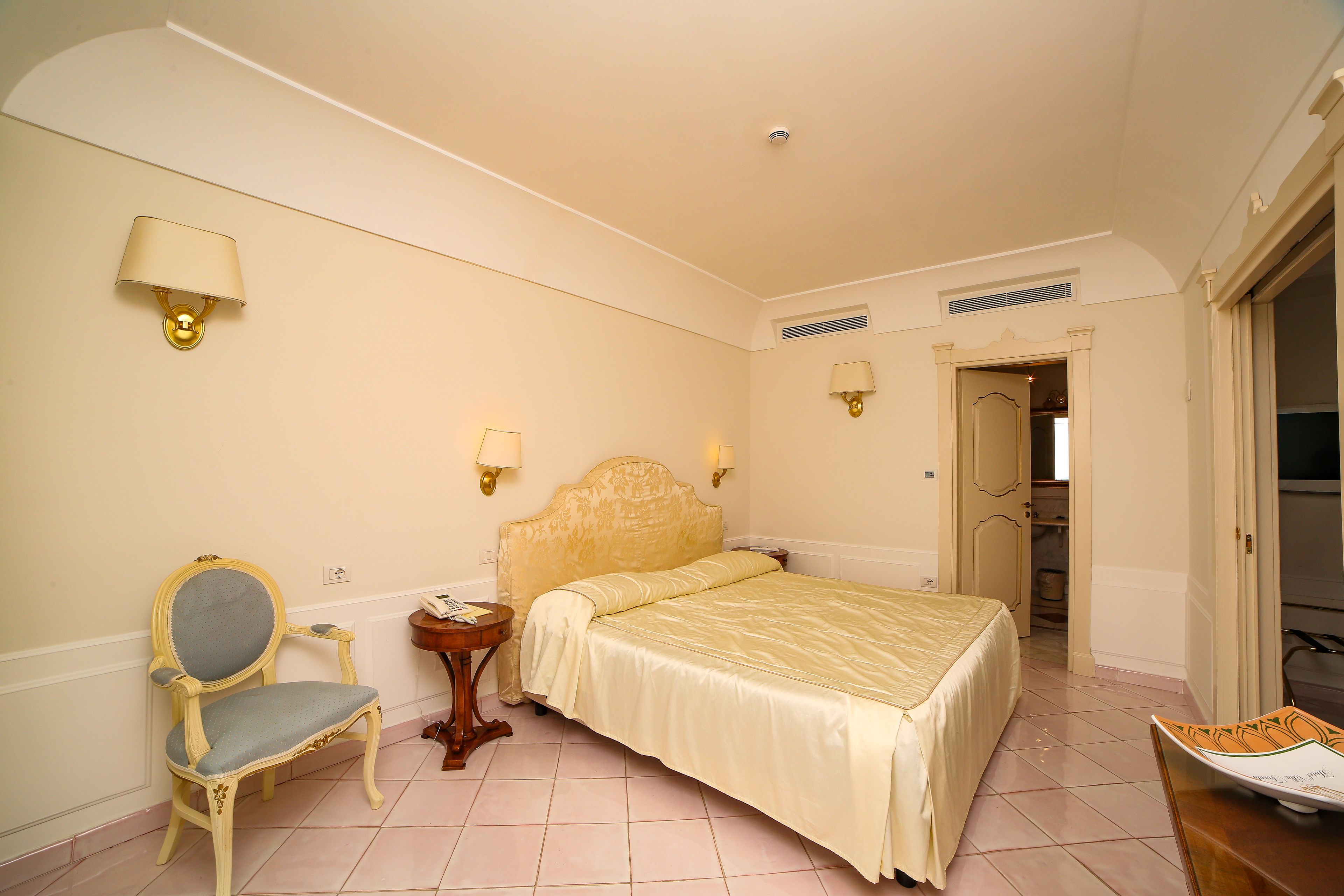A hotel room with a bed, a chair, a bedside table, and two wall-mounted lamps.
