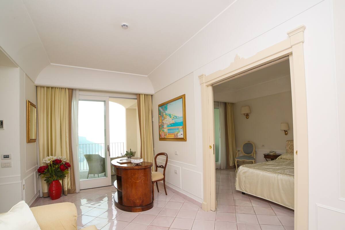 A hotel room with a balcony, a bed, a sitting area with a sofa, a wooden side table, and a painting on the wall.