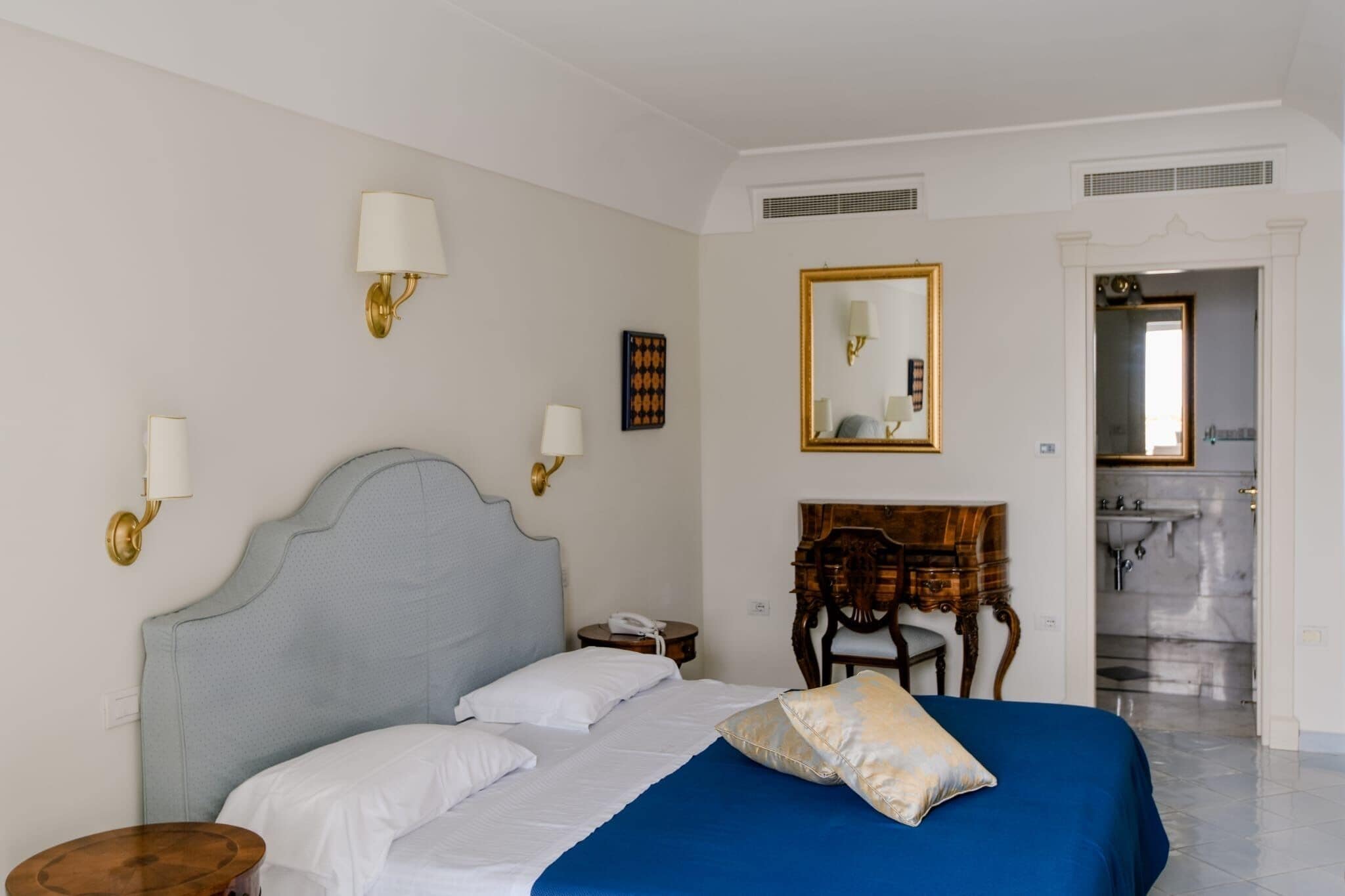 A hotel room with a bed, bedside lamps, a mirror, a wooden dresser, and a bathroom visible through an open door.