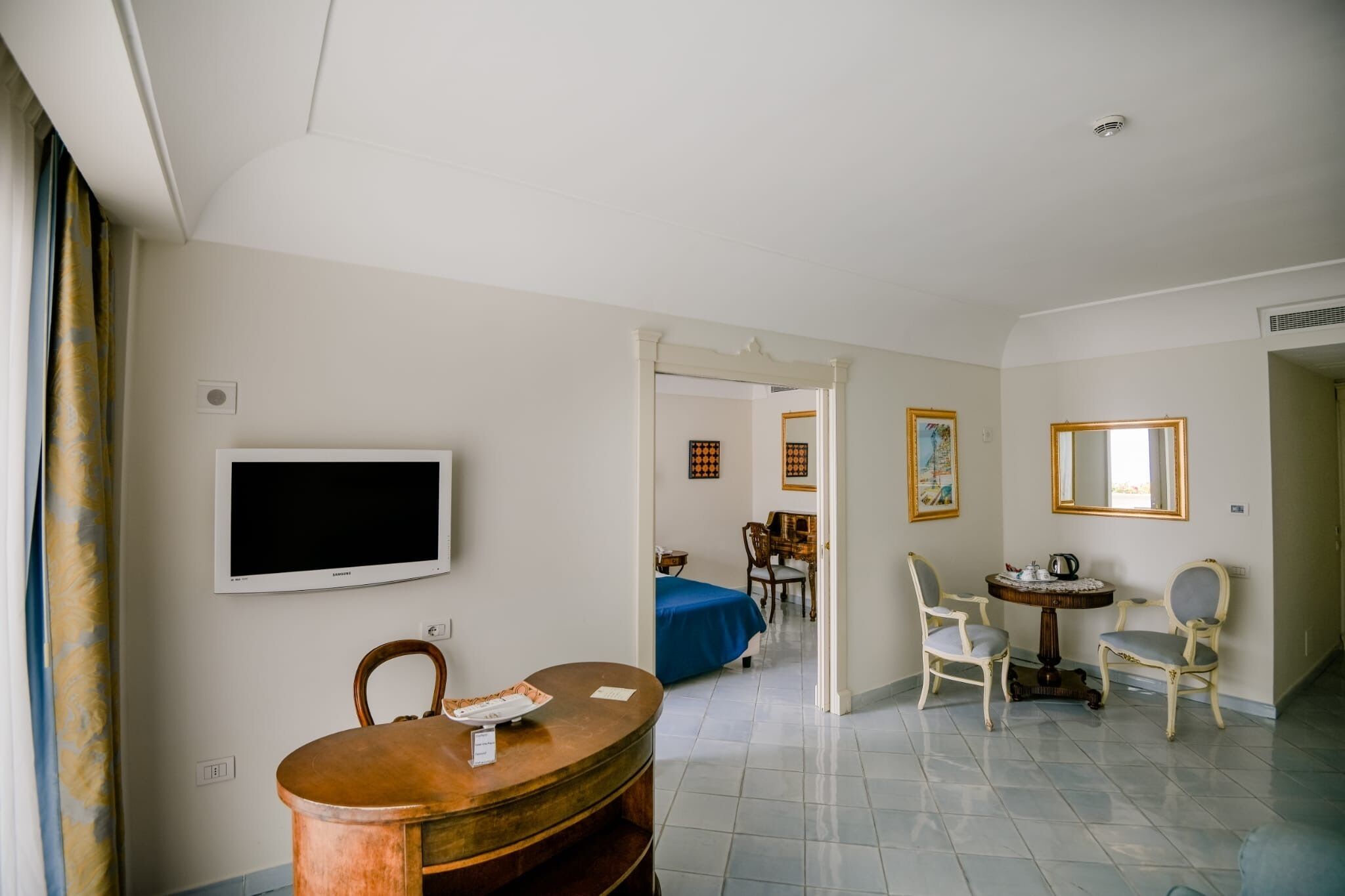 A room with a television, a dining table with chairs, a bed with a blue cover, and a small round table with chairs.