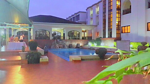 Un patio de hotel con piscina, mobiliario exterior y una zona cubierta con mesas y sillas.