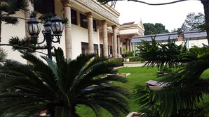 Un edificio con columnas y un paseo techado, rodeado de exuberante follaje y plantas en macetas.